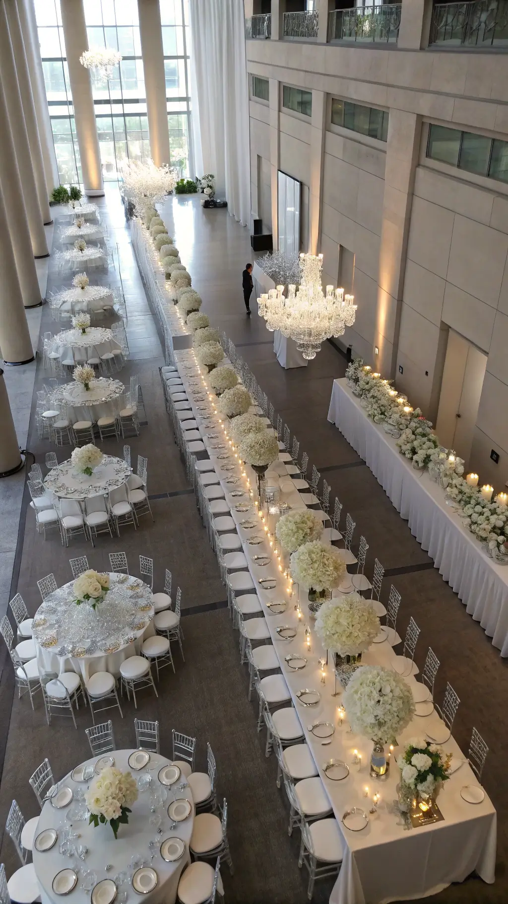 Aerial view of an elegant ceremony space with white-themed tablescapes; long and round tables feature white floral arrangements on clear risers, mercury glass votives, and crystal installations above; ghost chairs blend into the scene with natural light casting shadows on pewter flooring.