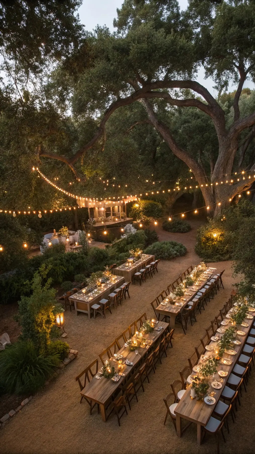 Aerial view of a garden wedding reception at twilight with U-shaped wooden tables under oak trees adorned with fairy lights. Tables are decorated with eucalyptus and rose garlands, brass vases, and votive candles. Cross-back chairs and vintage place settings complete the elegant scene, with market lights overhead twinkling like stars.