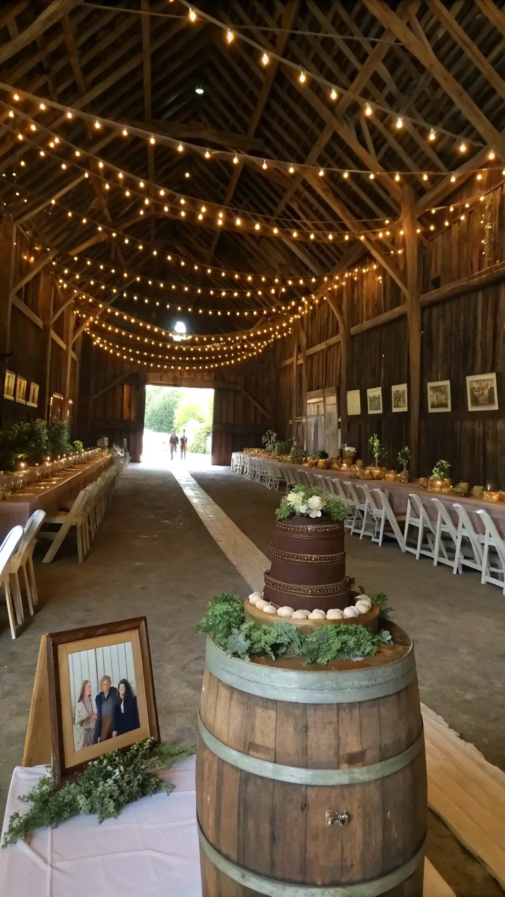 A warmly lit historic barn interior with cafe lights, showcasing a rustic chocolate peanut butter cake on a wine barrel, surrounded by wooden chairs and harvest tables with flowing runners and potted herbs; vintage couple photographs in antique frames enhance the cozy, nostalgic ambiance. 50th Wedding Anniversary Chocolate Cake