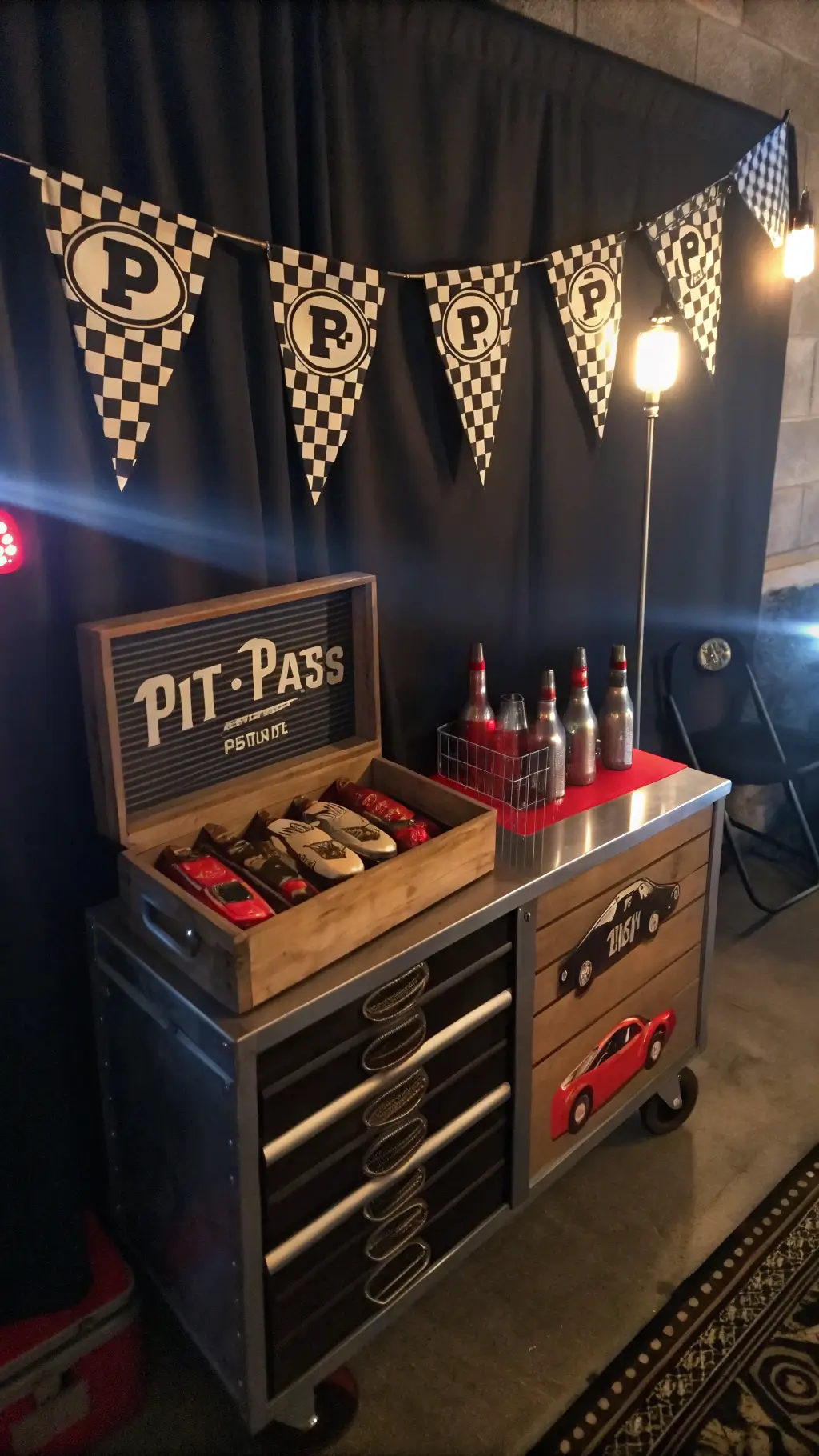 An intimate tabletop vignette featuring a 'pit pass' favor station under moody artificial lighting. Custom leather-look pit crew badges are displayed on a vintage tool cart next to chrome-finished boxes holding mini wooden race cars. Checkered fabric bunting drapes the setup, enhanced by LED strip lighting casting dramatic shadows. Racing gloves and branded water bottles are precisely arranged, with a fog machine adding atmosphere. Car Birthday Party