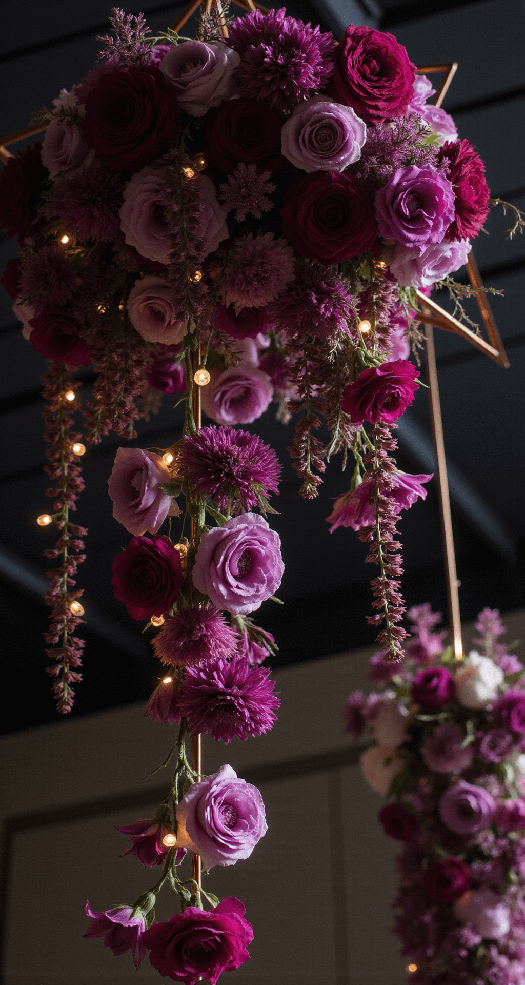 A dramatic close-up of a suspended floral installation featuring deep magenta and purple blooms cascading from copper geometric frames, with smooth calla lilies and ruffled garden roses, twinkling fairy lights in between, captured from below against a dark ceiling.