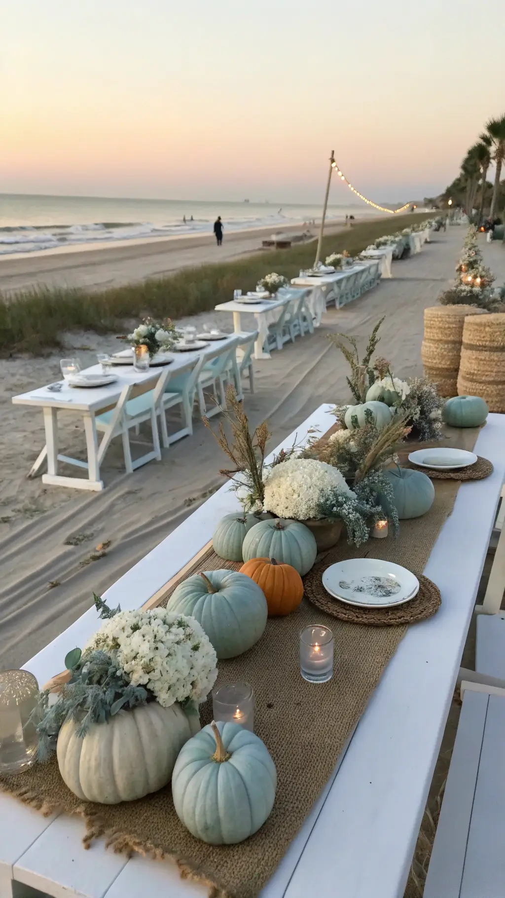 A scenic beachfront celebration at sunset showcasing coastal-themed pumpkin displays with whitewashed farm tables adorned with sea glass, driftwood, and blue-grey pumpkins, alongside white hydrangeas, dusty miller, and grasses; complemented by linen runners, rattan chargers, and shell accents, with the ocean horizon visible in the background.