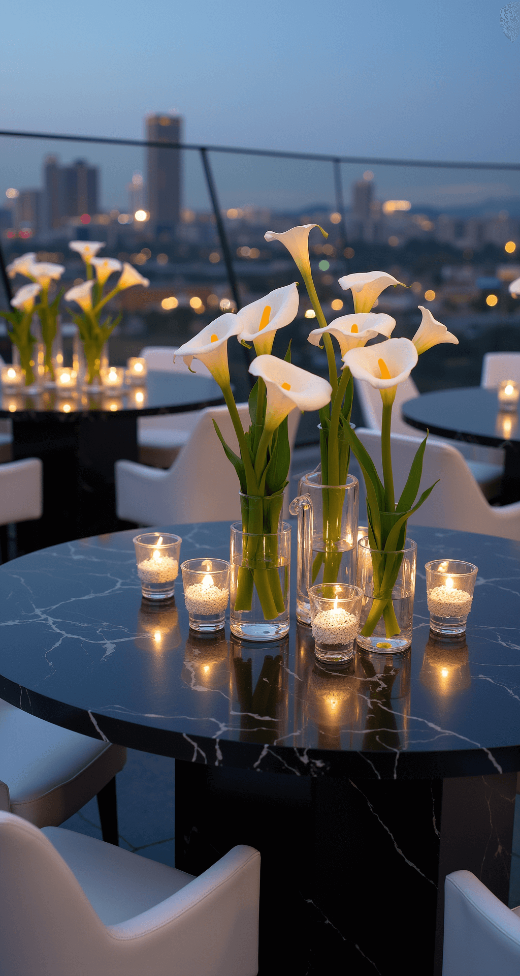 Modern rooftop venue at twilight featuring minimalist white calla lily arrangements in tall geometric vases atop black marble tables, with city lights twinkling in the background and uplighting enhancing each centerpiece, complemented by white leather seating and chrome accents, captured from an elevated angle.