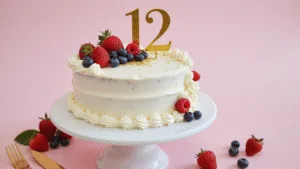 Photorealistic overhead shot of an elegant half-moon birthday cake with white buttercream frosting, topped with fresh raspberries, blueberries, and metallic gold sprinkles, featuring a gold "½" topper on a white ceramic stand against a soft pastel pink backdrop with bokeh effect.