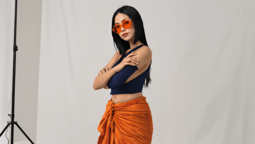A confident woman with long straight black hair poses against a clean white backdrop, wearing a navy cropped tank top and a burnt orange wrap sarong, accessorized with orange-tinted sunglasses and nude flats, showcasing an elegant and sophisticated anime-inspired fashion style.