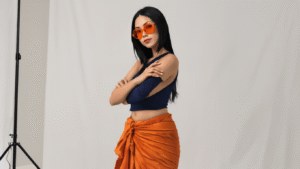 A confident woman with long straight black hair poses against a clean white backdrop, wearing a navy cropped tank top and a burnt orange wrap sarong, accessorized with orange-tinted sunglasses and nude flats, showcasing an elegant and sophisticated anime-inspired fashion style.