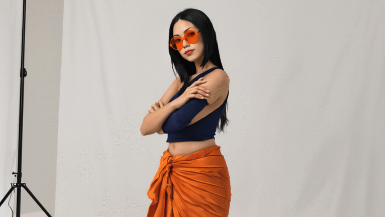 A confident woman with long straight black hair poses against a clean white backdrop, wearing a navy cropped tank top and a burnt orange wrap sarong, accessorized with orange-tinted sunglasses and nude flats, showcasing an elegant and sophisticated anime-inspired fashion style.