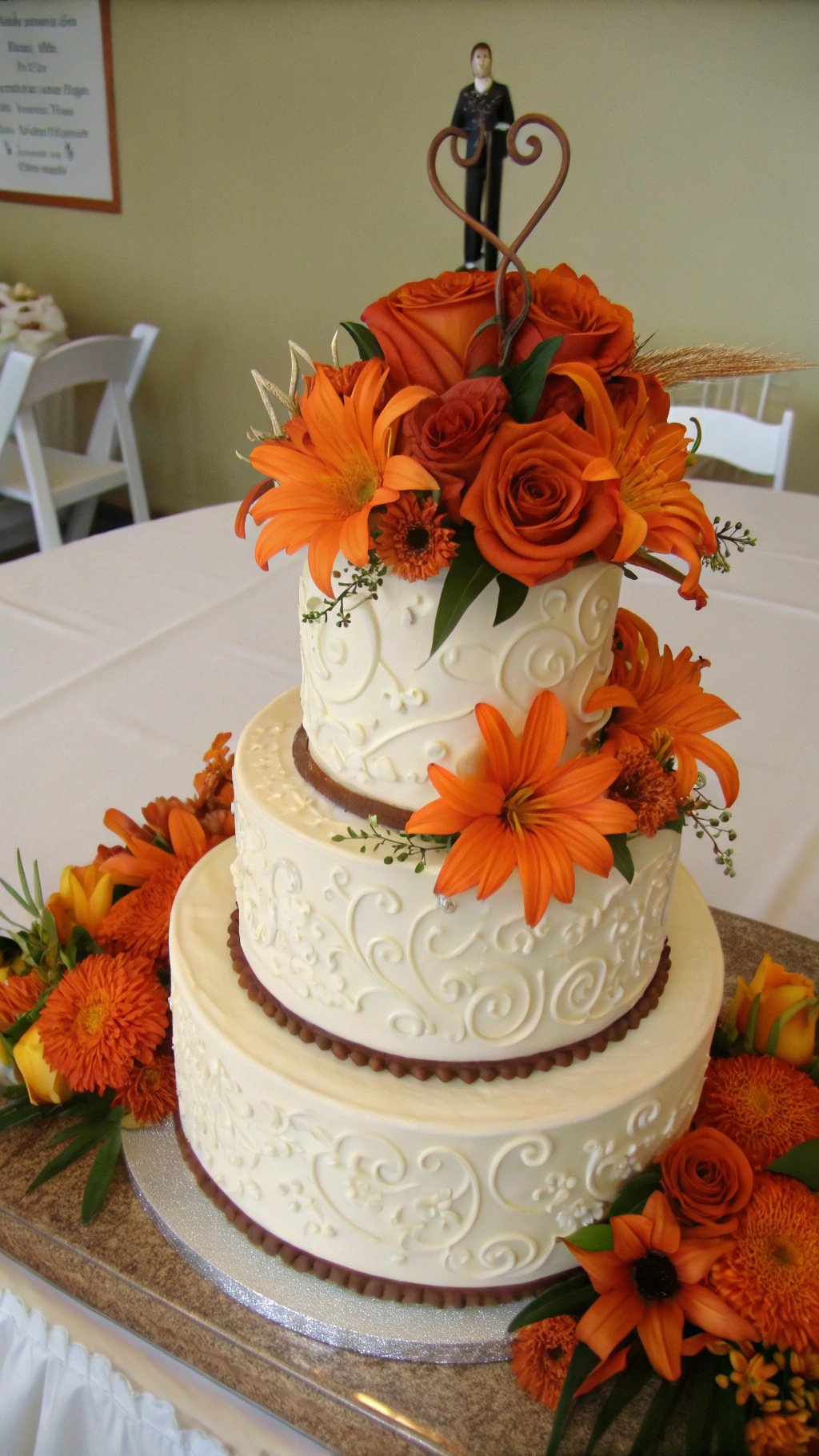 A three-tier wedding cake decorated with orange flowers and a couple figurine on top.