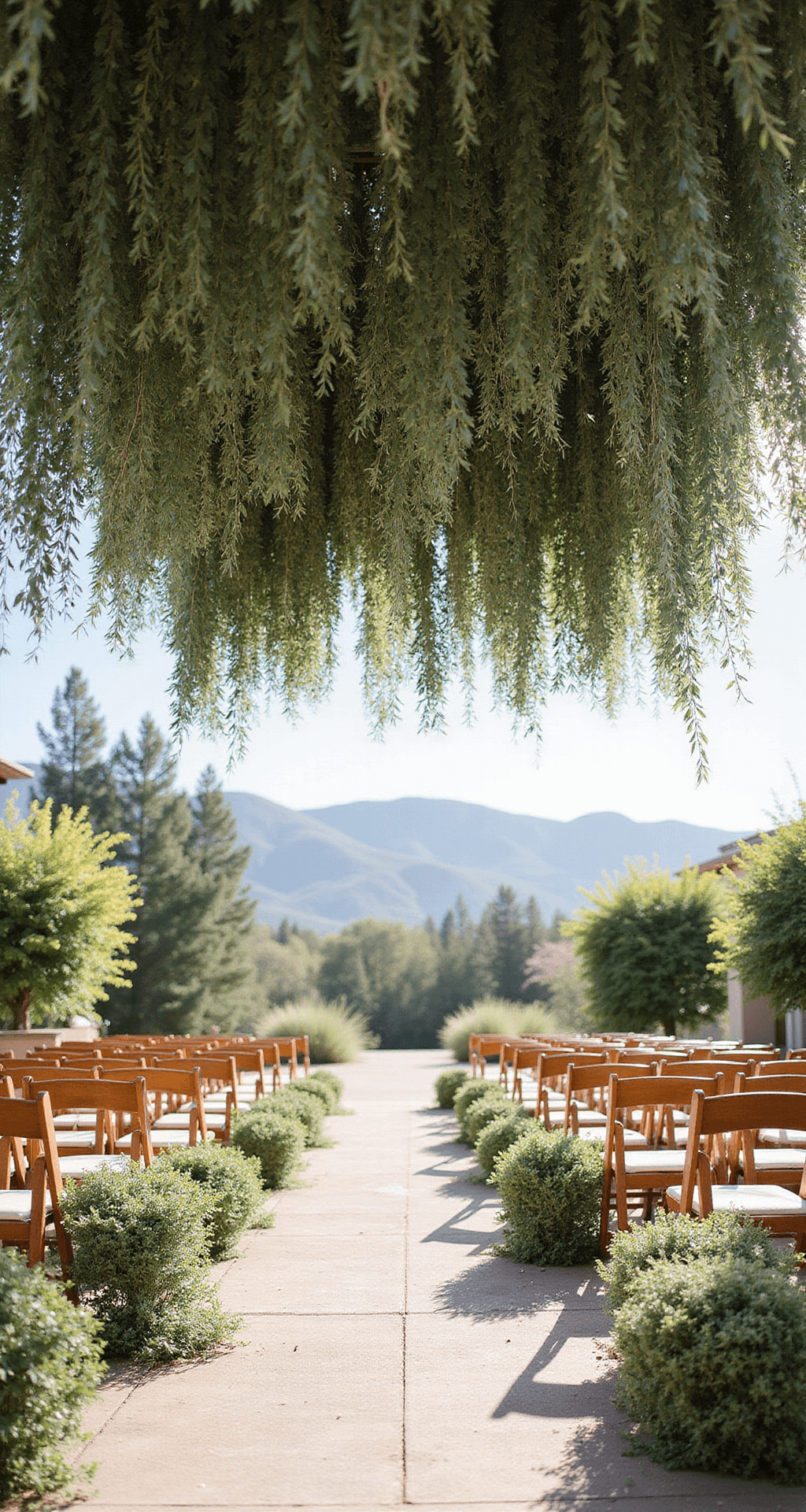 A breathtaking outdoor ceremony space adorned with suspended garlands of eucalyptus and olive branches, bathed in soft sunlight, featuring wooden chairs with minimal fabric accents, set against a romantic garden backdrop with distant mountains, captured from an overhead perspective.
