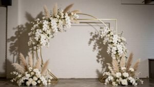 A dramatic statement wedding backdrop featuring an asymmetrical metal arch with cascading white orchids and pampas grass against a modern white brick wall